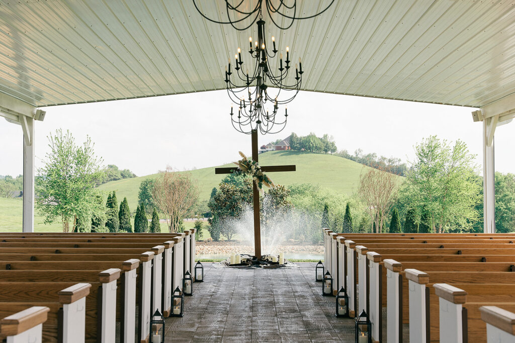 A view down the aisle of the outdoor wedding ceremony space with wooden pews and cross under black chandeliers at The White Dove Barn