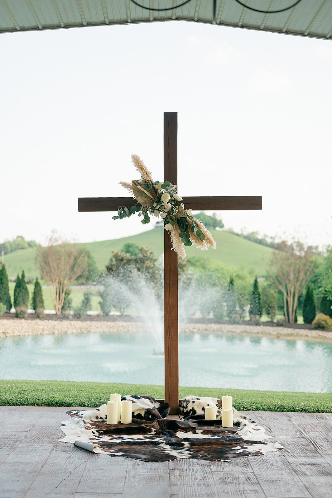 A view of a wooden cross with florals and candles in front of the fountain at The White Dove Barn