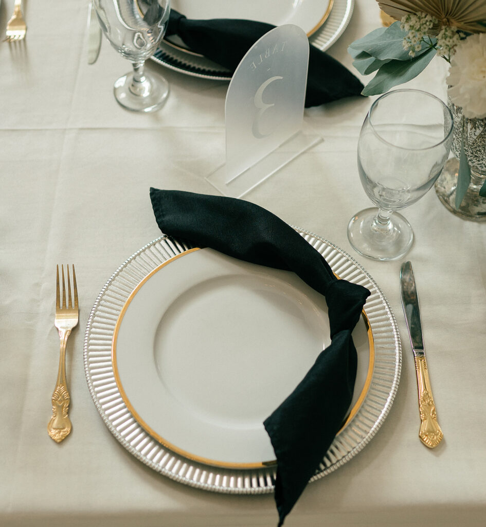 A black napkin sits on a gold rimmed charger plate with gold silverware on white linen