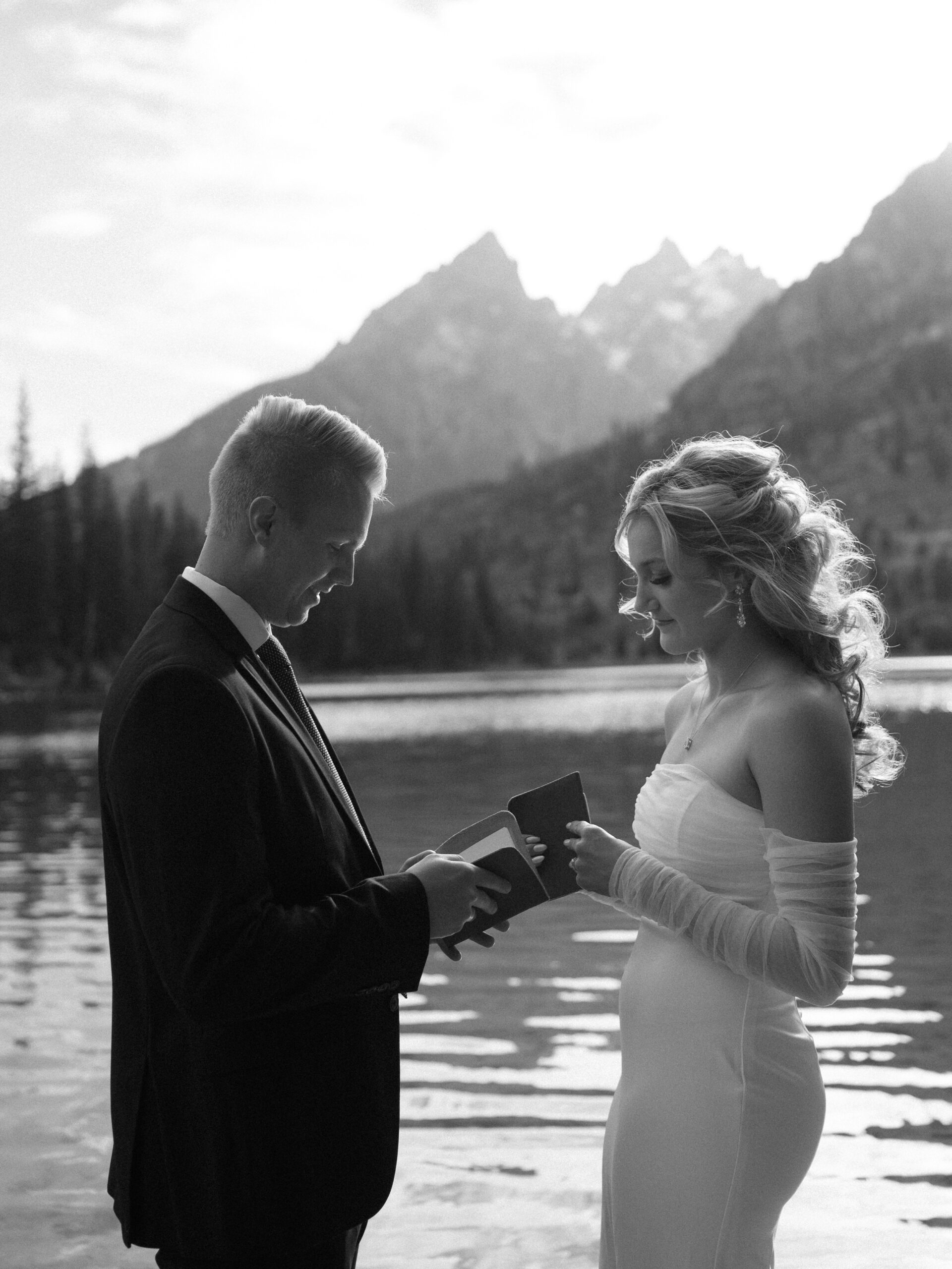 A bride and groom in black and white exchange vows lakeside in the mountains