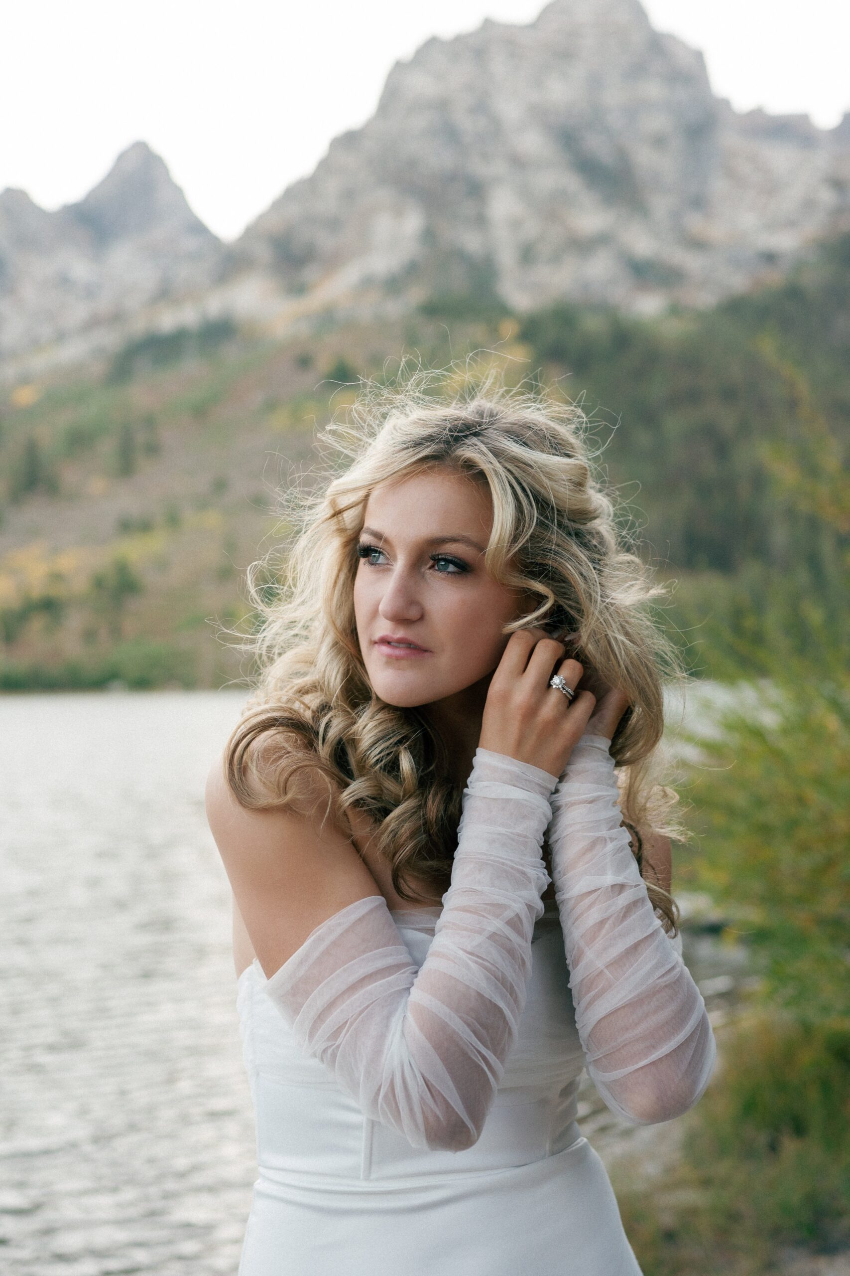 A bride adjusts her earrings while standing lakeside at one of the Jackson Hole Wedding venues