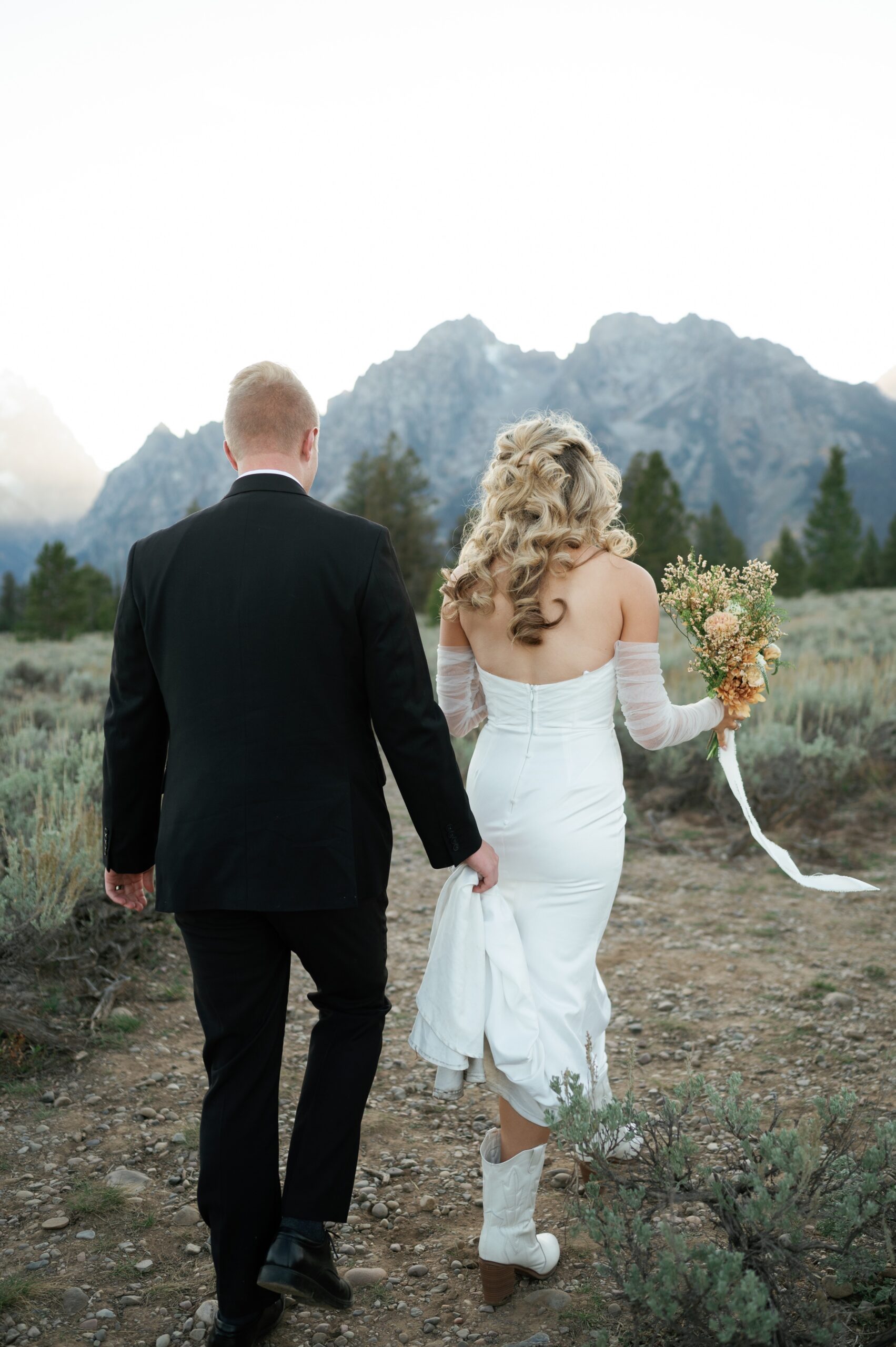 A groom holds his bride's train as their hike in a mountain trail at sunset