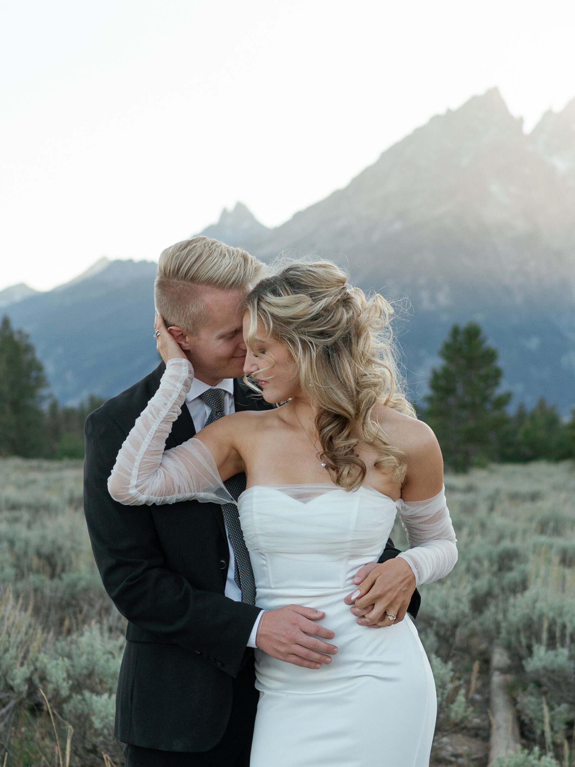 A groom smiles while nuzzling his bride in a mountain meadow