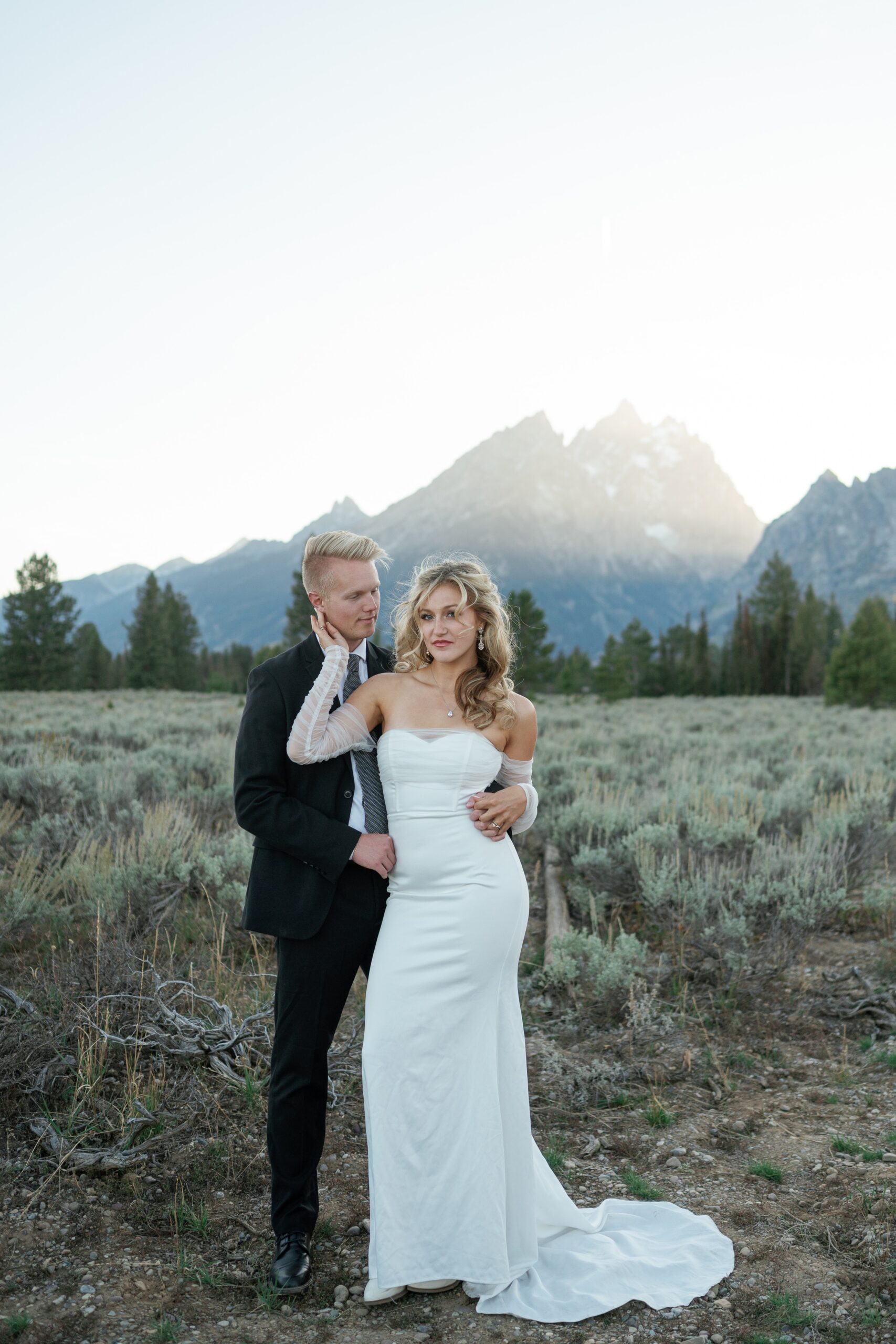 A groom stands with his bride as she holds his neck in a mountain meadow trail at sunset