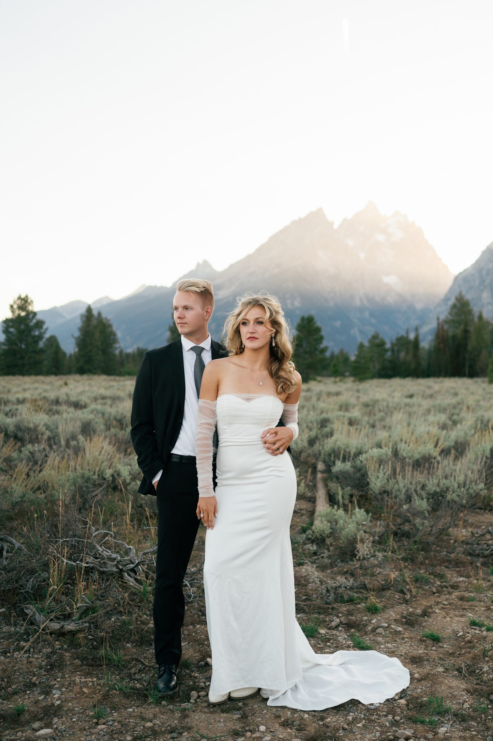 A bride and groom stand together gazing out into the meadow at sunset