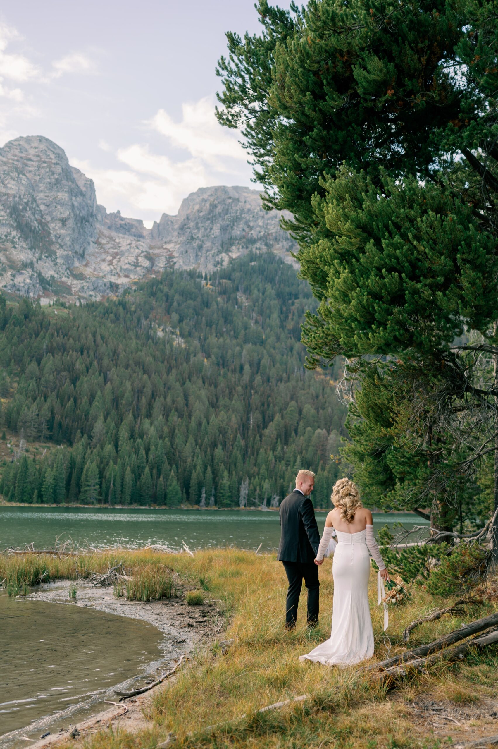 A groom leads his bride along a lake trail in the mountains at one of the Jackson Hole Wedding venues