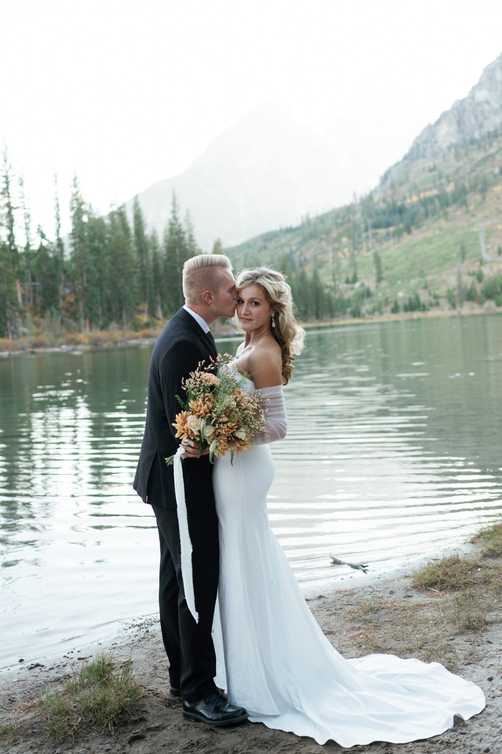 A bride is kissed by her groom while standing on the dge of an alpine lake