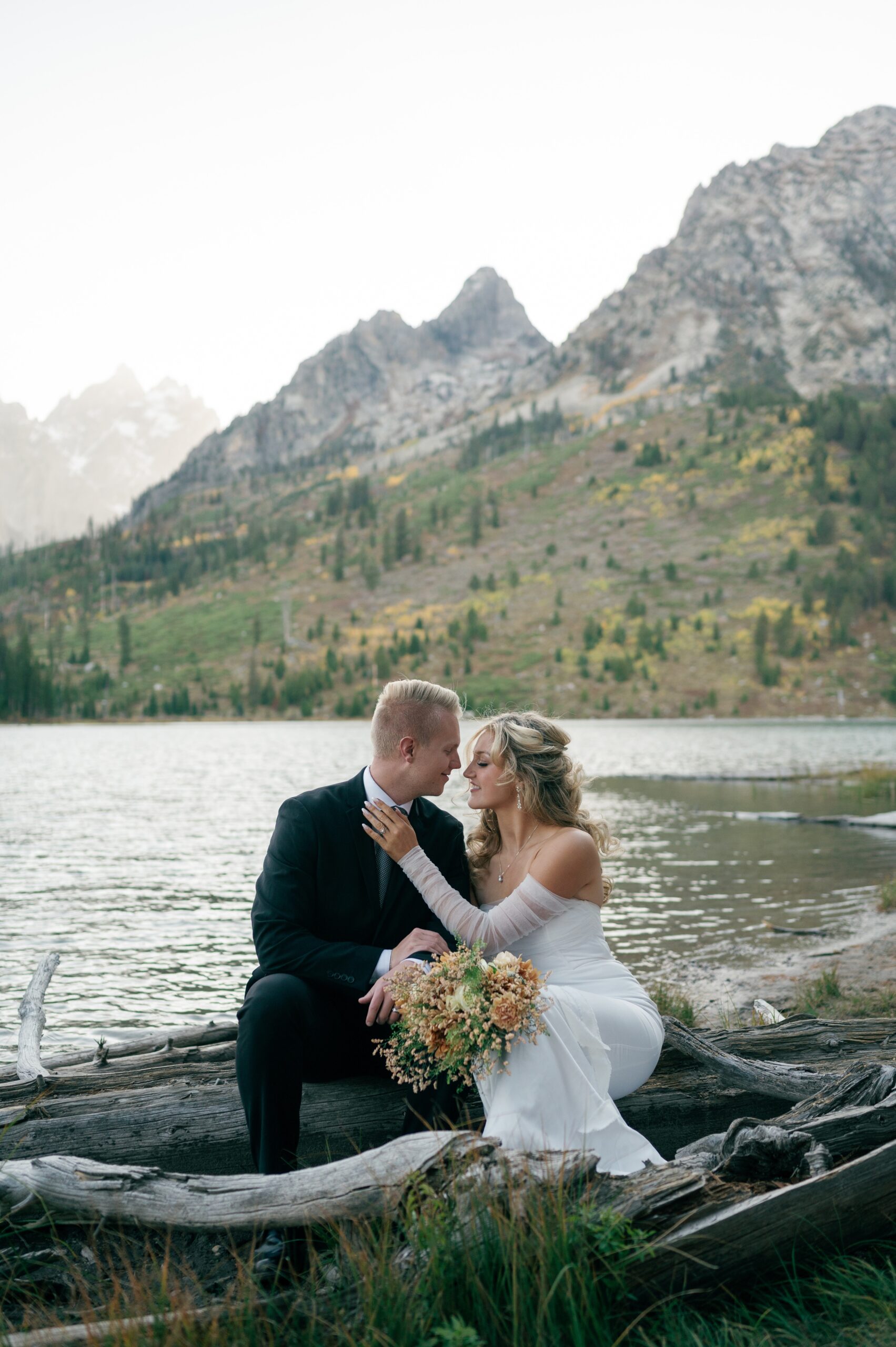 Newlyweds lean in for a kiss while sitting on driftwood by an alpine lake