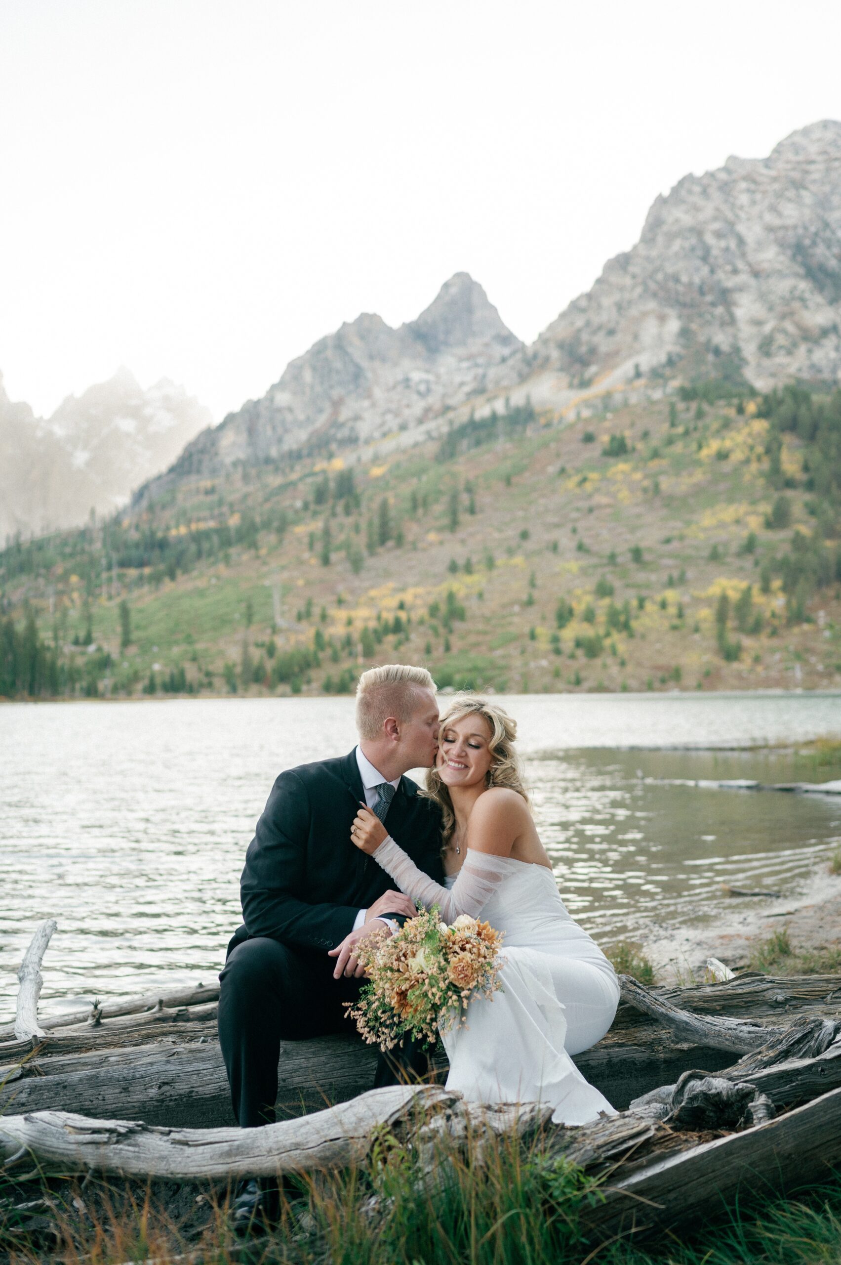 A bride smiles as her groom kisses her on some driftwood by a lake at sunset