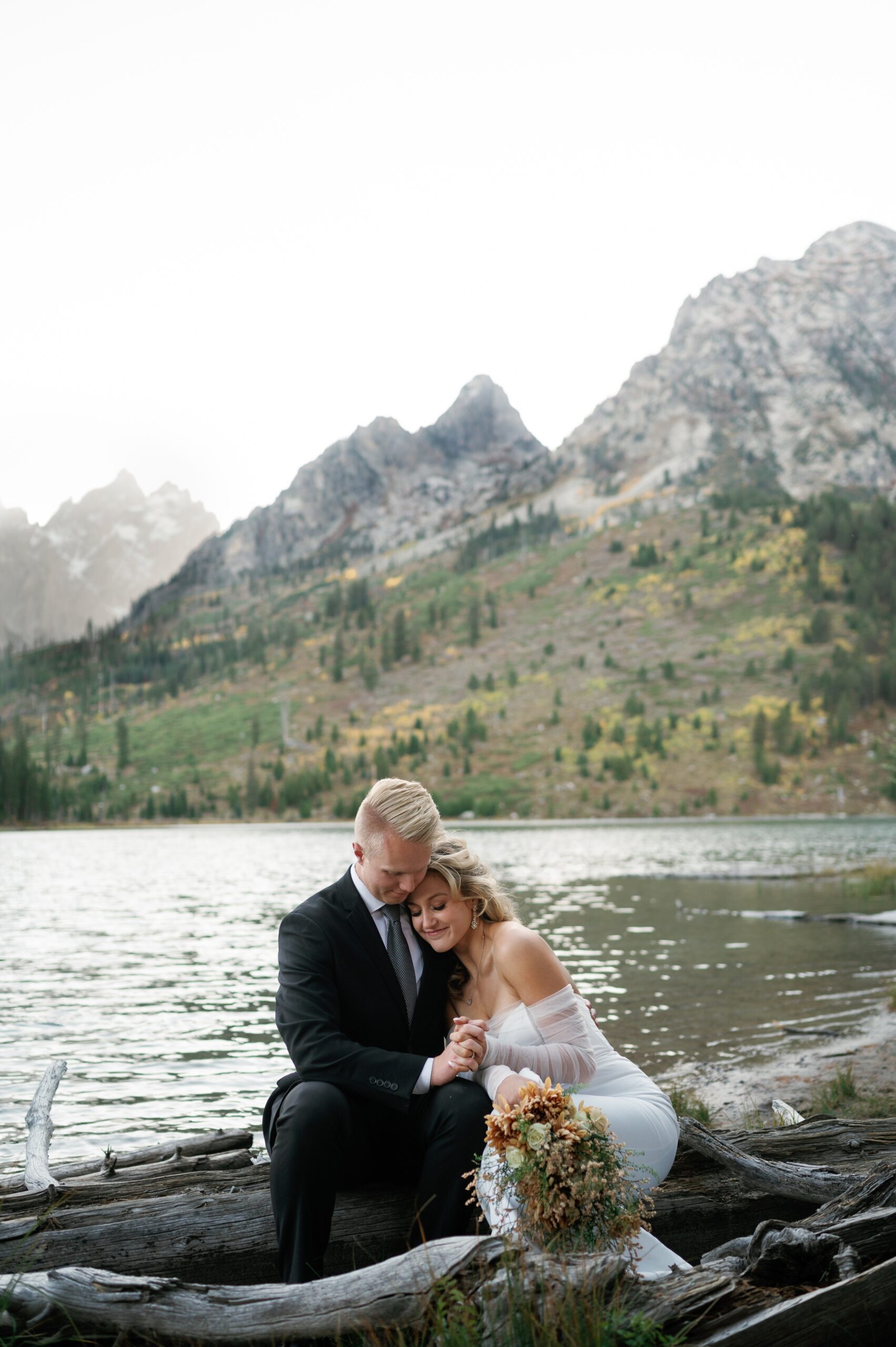 Newlyweds snuggle on driftwood on a lake in the mountains at one of the Jackson Hole Wedding venues