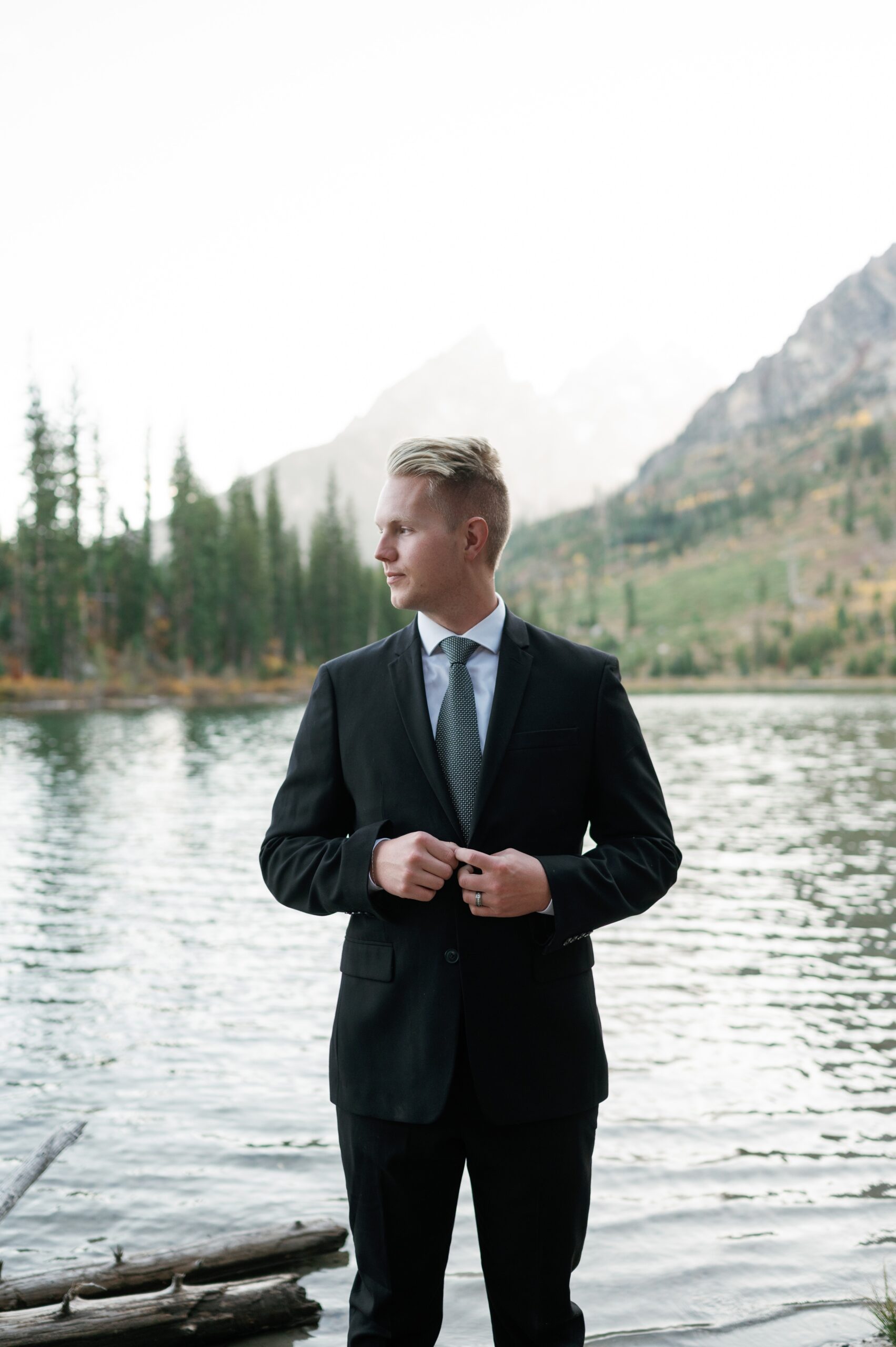 A groom adjusts his jacket button while looking over his shoulder at an alpine lake at one of the Jackson Hole Wedding venues