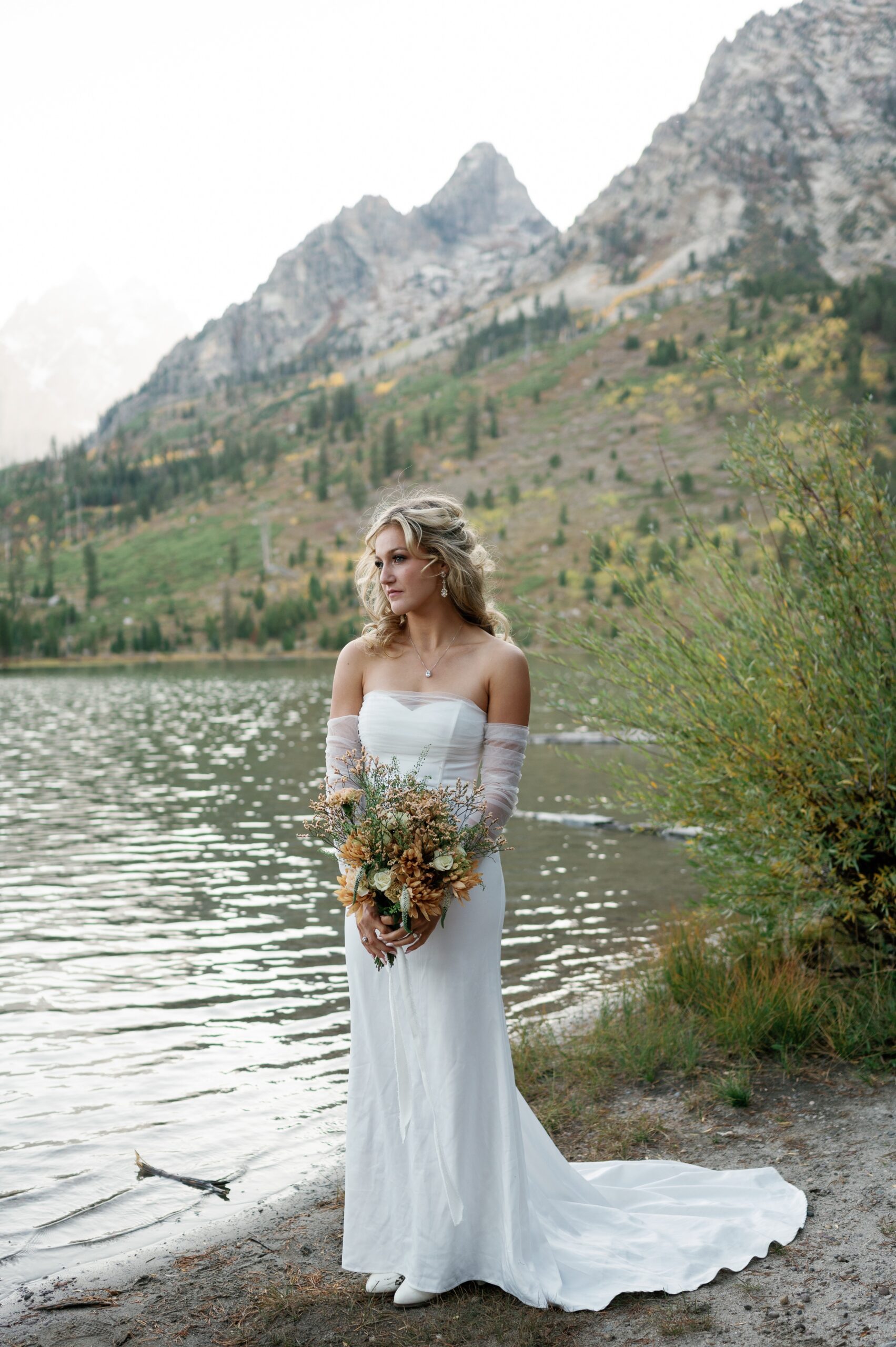 A bride holds her bouquet while standing on the edge of a lake at sunset in the mountains